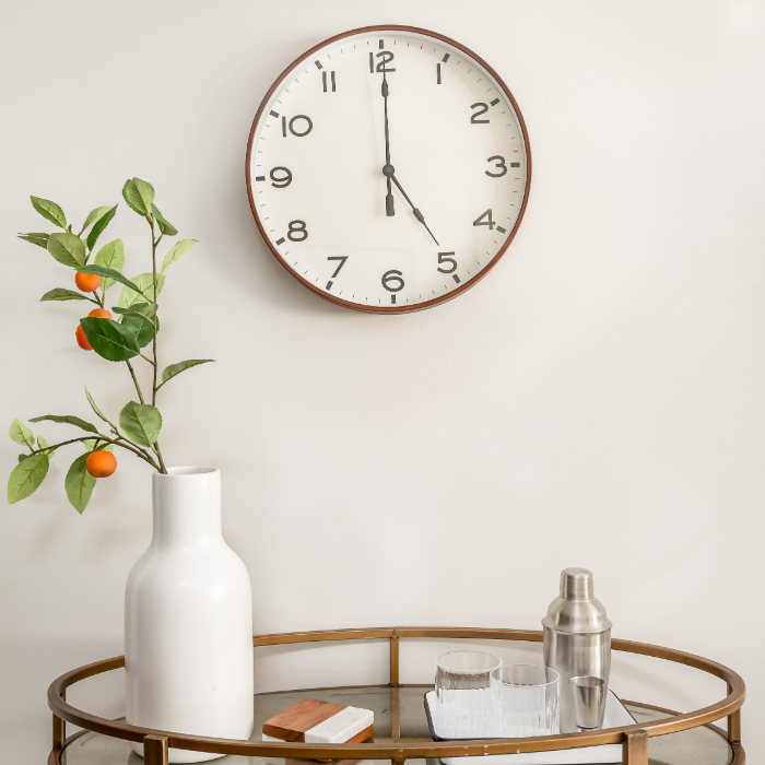 Relógio de parede com design simples e números pretos em ambiente decorado com bandeja, copos e vaso com galho de frutas laranjas