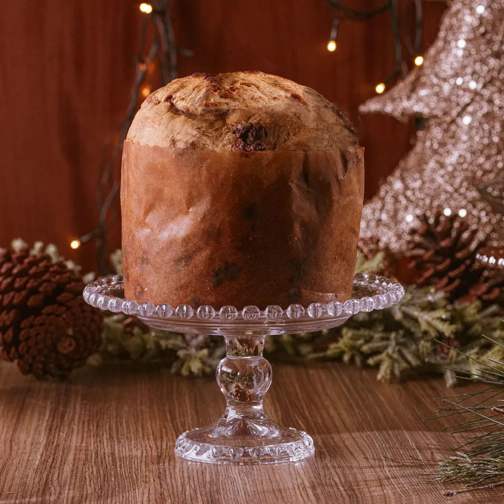 Boleira de vidro transparente com pé elevado e bordas decoradas em relevo, exibindo panetone em uma mesa de madeira com pinhas, luzes e decoração natalina.