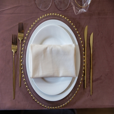 Sousplat transparente com borda de esferas douradas, pratos brancos, guardanapo de tecido e talheres dourados em uma mesa posta refinada.