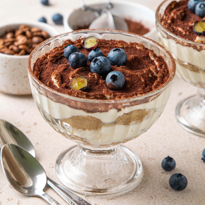 Sobremesa servida em uma taça de vidro transparente com base de creme claro, camadas de biscoito embebido e cobertura de cacau em pó, finalizada com mirtilos frescos. Ao fundo, utensílios de cozinha e grãos de café complementam a cena.